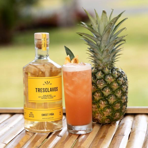Bottle and glass of Tres Clavos Sweet Piña with pineapple on wooden surface.