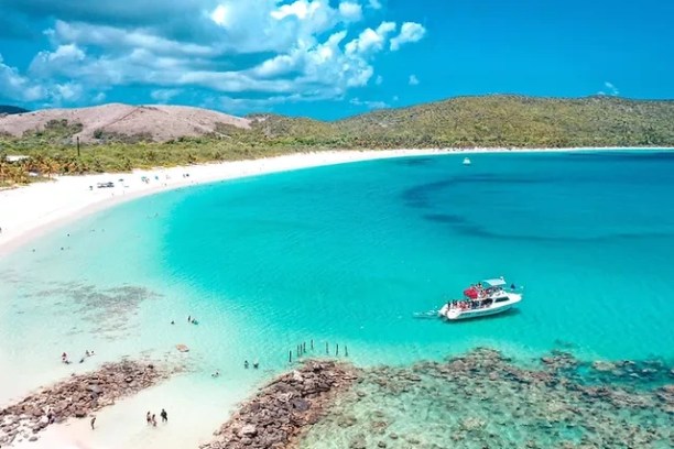 Boat in clear blue water near a beach with few people and distant hills.