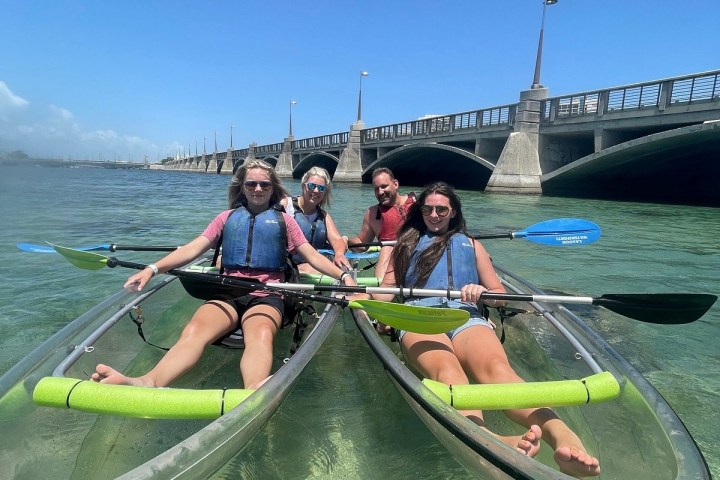 Four people in clear kayaks near a bridge, smiling on a sunny day.