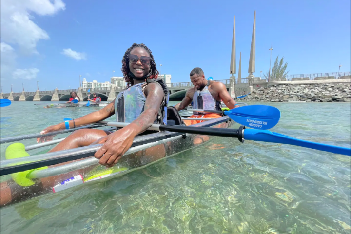 Smiling people kayaking on clear water with distant buildings.