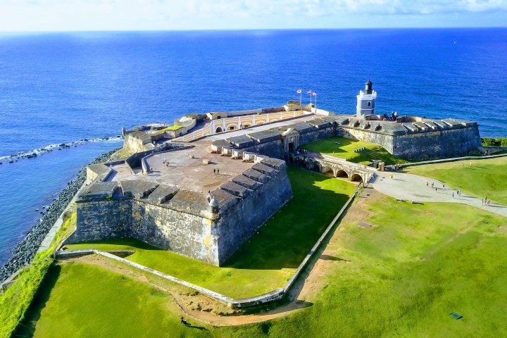 Aerial view of a historic coastal fortress with a lighthouse, surrounded by grass and overlooking the ocean.