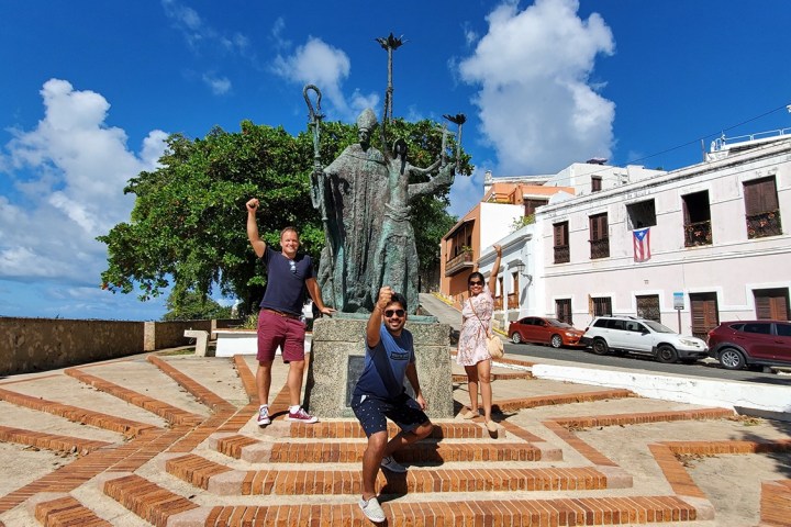 Three people posing by a historic statue in a sunny plaza.
