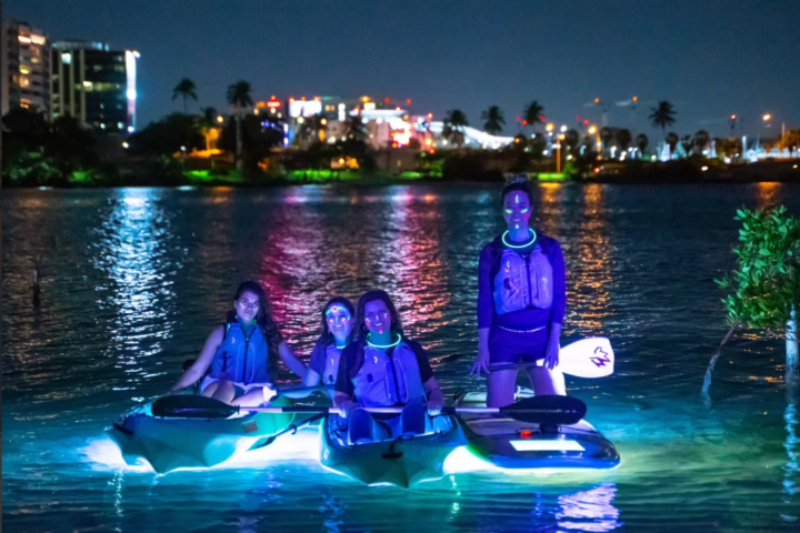 Four people kayaking on a glowing river at night with city lights in the background.