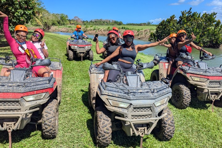 Group of people on ATVs, wearing helmets, posing energetically on a grassy path by a river.