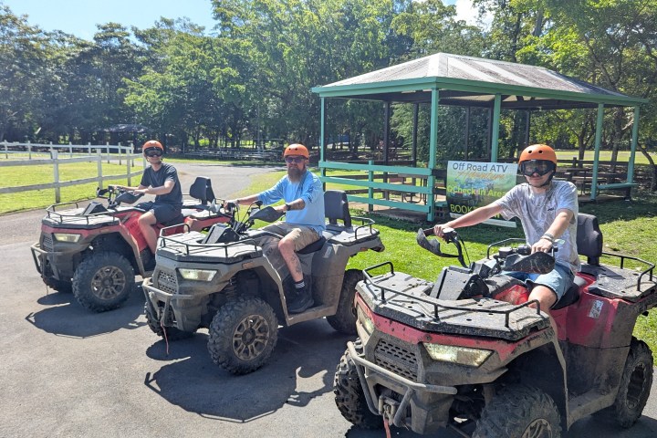 Three people on ATVs wearing helmets near a pavilion in a park.