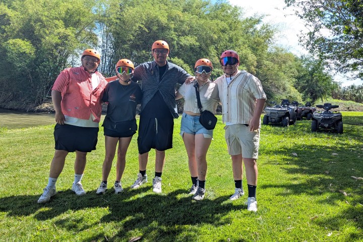 Five people in helmets posing outdoors near trees and quad bikes.