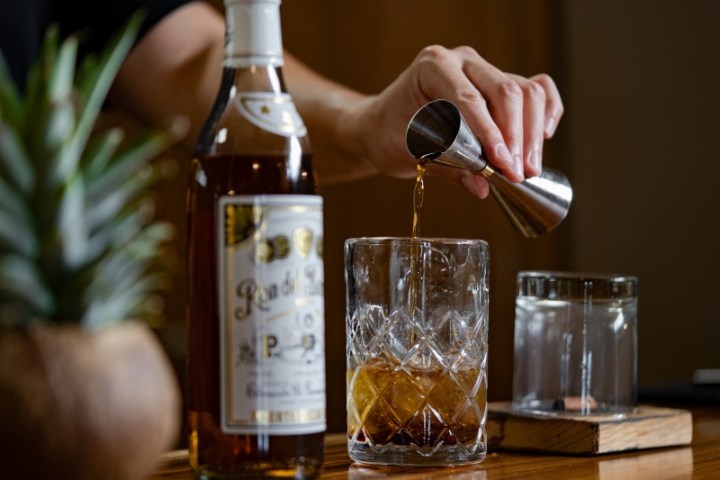 Person pouring liquor from jigger into glass next to a bottle on a wooden table.