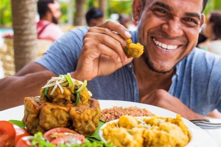 Man smiling, holding food near plate with salad, greens, and a creamy dish outdoors.