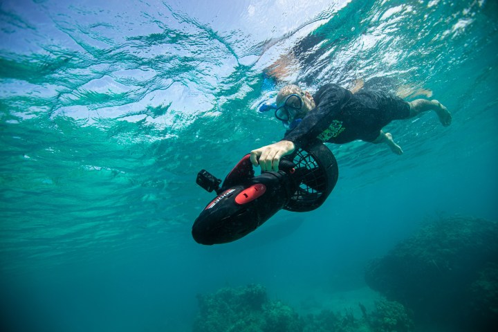 Person snorkeling underwater holding a black scooter in clear blue water.
